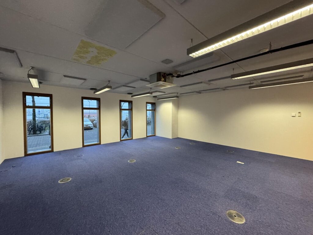 Empty office space on De Ruijterkade with blue carpet, white walls, and large windows facing the street.